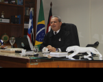 Imagem com fundo branco e bandeiras do Maranhão, Brasil e da Justiça do Trabalho, tendo ao centro o presidente do TRT no Maranhão, desembargador Carvalho Neto, sentado em seu gabinete, vestindo terno escuro.