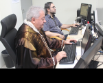 Juiz Veloso Sobrinho veste toga de cor marrom e está sentado ao lado do chefe de audiência Daniel Dantas que veste camisa listrada. Ambos estão na sala de audiências e em frente à tela de computadores.