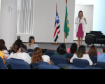 Foto de pessoas sentadas e de uma mulher em pé no palco de um auditório, próximo às bandeiras do Brasil e do Maranhão e de uma poltrona preta.
