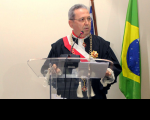 Foto de um homem usando toga, na tribuna. Ao fundo, bandeira do Brasil .