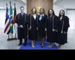 Foto de quatro mulheres e um homem de pé, sorrindo e posando para a câmera. Atrás deles, parede branca com recorte amadeirado onde se vê o brasão da República em dourado. À esquerda, veem-se bandeiras hasteadas.