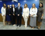 Foto de um homem usando terno escuro ladeado por seis mulheres. Todos estão de pé e posam para a câmera, sorrindo. Atrás deles, parede azul e mesa de madeira.