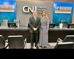 Foto de um homem usando terno escuro e gravata azul ao lado de uma mulher usando vestido estampado em preto, branco e azul Ambos estão de pé, posando para a câmera. Atrás deles, vê-se mesa escura com cadeiras cinza e parede em tons de cinza e azul com a logomarca do CNJ centralizada. Nas laterais da parede, ilustração com o nome do Encontro Nacional do Poder Judiciário.