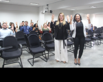 Foto mostra duas mulheres em pé, posicionadas em frente às cadeiras de um auditório. Elas vestem roupas sociais nas cores preto e branco. Ao fundo pessoas diversas sentadas. Todas estão com as mãos erguidas para cima, fazendo um sinal em Libras. 