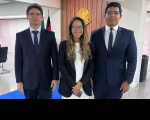 Foto mostra no primeiro plano e no centro uma mulher usando roupas sociais nas cores branco e preto, ela tem cabelos longos, usa óculos. Ela está ladeada por dois homens que usam ternos escuros, com camisas brancas e gravatas no tom azul.
