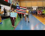 Uma pessoa segurando uma placa que identifica a delegação do TRT-16. Ao fundo, atletas seguram a bandeira do Maranhão
