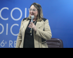 Foto de mulher loira, usando óculos e sorrindo enquanto fala ao microfone, que segura com a mão esquerda. Ela usa blusa preta e blaser bege com detalhes pretos. Atrás dela, vê-se parede azul com inscrição branca e espaldar de uma cadeira marrom.