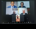 Foto de fundo escuro, mostrando print de uma tela de computador com cinco pessoas sorrindo e posando para a câmera durante realização de uma audiência virtual.