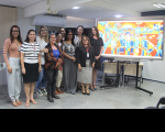 Foto de dez mulheres de pé e sorrindo para a câmera. À sua esquerda, tela de projeção mostrando quadro colorido em tons de laranja e azul.