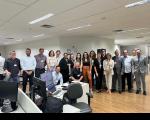Foto em um escritório com muitas pessoas em pé, sorrindo para a câmera. À frente, mesas com computadores e equipamentos.