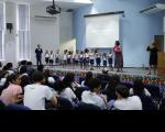 Foto de várias crianças e duas professoras em cima de um palco, ao fundo paredes claras. Em um plano mais baixo, várias pessoas sentadas em poltronas azuis.