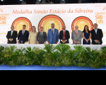  Foto mostra a presidente do TRT-16, desa. Márcia Andrea Farias posicionada na mesa de autoridades com outras personalidades. Na parte de cima, destaque para a frase: Medalha Simão Estácio da Silveira.