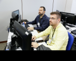 Imagens de dois homens brancos sentados. Ambos usam camisa social manga longa. Os dois estão utilizam computadores de mesa. Ao fundo uma parede branca e mais monitores.