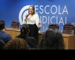 Foto mostra uma mulher em pé, diante de uma plateia. Ao fundo uma parede azul e a logo da Escola Judicial