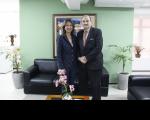 Foto mostra uma mulher e um homem. Ambos estão de pé. Ao fundo um ambiente de gabinete com sofá, quadros na paredes.