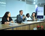 A imagem mostra quatro pessoas sentadas a uma mesa. Da esquerda para a direita, estão: uma mulher de cabelo preto; um homem de terno, usando um microfone; uma mulher negra, com cabelo curto e blusa clara; e uma mulher com cabelo preto. A mesa tem copos de água, microfones e papéis