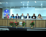 A imagem mostra uma sala com sete pessoas sentadas a uma mesa. No centro da mesa, um homem segura um microfone e lê um papel.