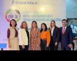 Foto de cinco mulheres e um homem de pé e sorrindo. Atrás deles, painel de cor clara com informações sobre o evento.