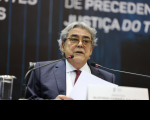 Foto de um homem claro, de cabelos grisalhos e usando óculos. Ele fala ao microfone enquanto segura papéis que lê.