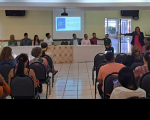 Foto de oito pessoas sentadas a uma mesa de frente para uma plateia. À direita, uma mulher de pé fala ao microfone, de frente para a plateia e apontando, com a mão direita, para tela de projeção, onde é exibida uma apresentação de slides. Plateia repleta de pessoas sentadas em cadeiras pretas.
