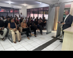 A foto mostra uma sala. Na parte direita, um homem em pé usa um terno escuro e camisa branca - sem gravata. Na esqueda vários homens e mulheres estão sentados. 