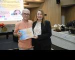 Foto mostra um homem com cabelos grisalhos e uma mulher branca no centro da imagem. Os dois seguram um certificado. Ao fundo um painel.