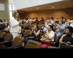 Pessoas sentadas em um auditório. Uma mulher branca se apresenta diante de todos.