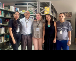 Foto mostra cinco pessoas em pé. Ao fundo prateleiras de alumínio com livros.