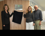 Foto mostra duas mulheres e um homem ao lado de uma placa de inauguração posicionada na parede. As duas mulheres 