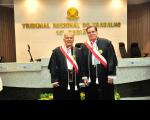 Foto mostra dois homens, dentro de um plenário. Ambos estão em pé e usam togas. Ao fundo, tem um brasão e o nome Tribunal Regional do Trabalho.