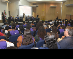 Foto mostra várioas pessoas sentadas em um auditório. Ao fundo, outras seis pessoas estão posicionadas no dispositivo de honra.