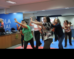 Foto mostra um grupo de mulheres, realizando exercícios de defesa pessoal. O grupo está dentro de um auditório