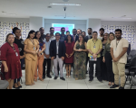 Foto mostra um grupo de pessoas em pé, são homens e mulheres. Todos estão em um auditório