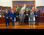 Foto mostra sete pessoas em pé, uma ao lado da outra. Ao fundo posicionado na parede um grande quadro com a imagem da Deus símbolo do Direito.
