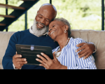 Foto de um homem e mulher negros e idosos abraçados e sorrindo, olhando a tela de um tablet, sentados em um sofá.