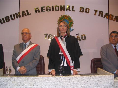 Mulher de toga, entre dois homens de terno, todos em pé, atrás de uma mesa de honra