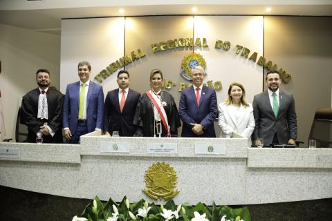 Mulher de toga, entre dois homens de terno, todos em pé, atrás de uma mesa de honra