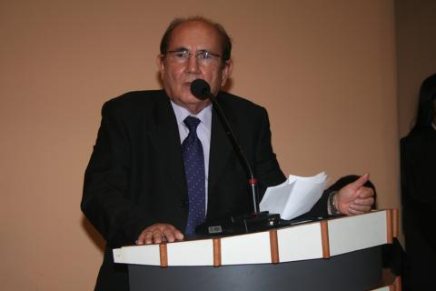 Homem de terno, em pé, em frente a um tribuna discursando