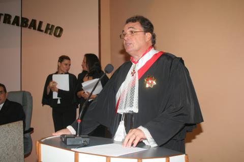 homem de toga em pé com uma faixa branca e vermelha discursando atrás de uma tribuna