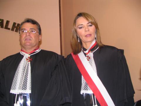 Mulher ao lado de um homem, ambos de toga, ela está com uma faixa vermelha e branca. .