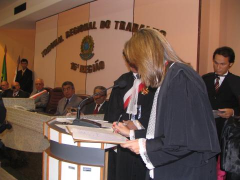Mulher de toga, atrás de uma tribuna, assinando o ato de posse.