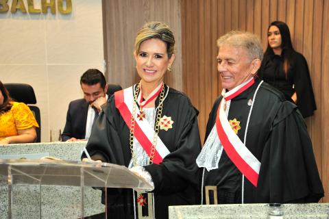 Mulher ao lado de um homem, ambos de toga, atrás de uma tribuna, a mulher está assinando o ato de posse .