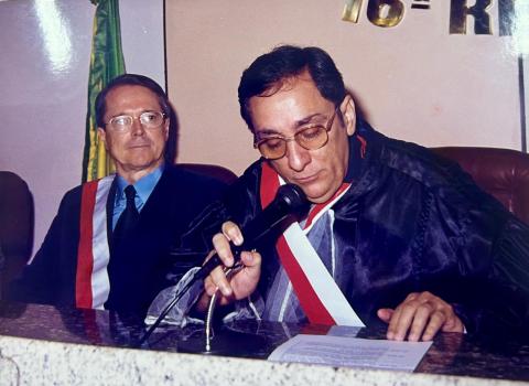 Homem de toga, sentado atrás de uma mesa,  falando em um microfone e segurando um papel