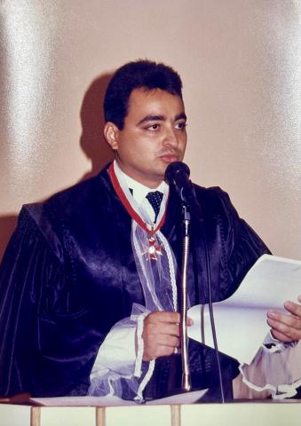 Homem de toga, em pé, em frente a um tribuna discursando