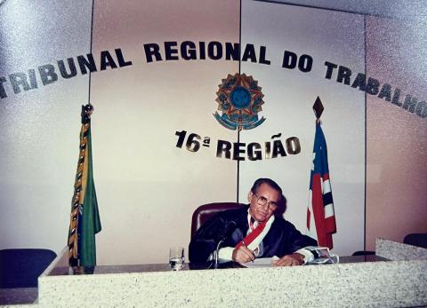 Homem de toga, sentado atrás de uma mesa,  assinando um papel