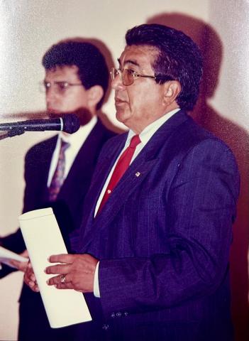 Homem de terno, em pé, em frente a um tribuna discursando