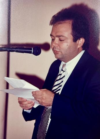Homem de terno, em pé, em frente a um tribuna discursando